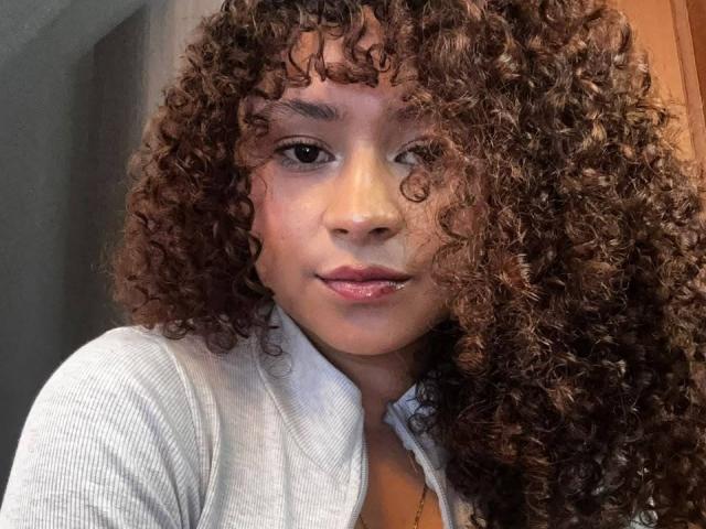 SofiaPassionne has curly brown hair, a light brown skin tone, and is wearing a grey ribbed shirt. She's winking with pink lips slightly parted.