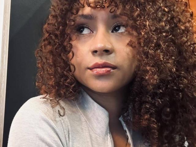 SofiaPassionne has curly brown hair, medium brown skin, and full pink lips with a small cut on her lower lip. She's wearing a gray shirt, looking to the side.