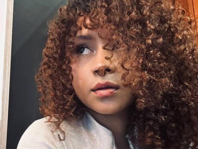 SofiaPassionne has curly brown hair covering one eye, light brown skin, and shiny pink lips. She's wearing a grey shirt, looking slightly to the right.