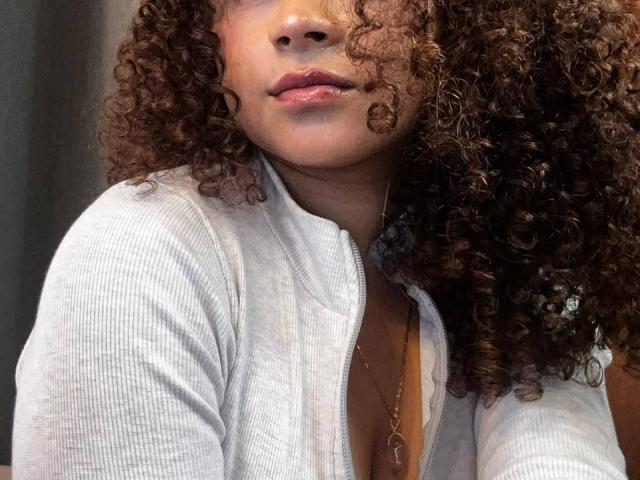 SofiaPassionne has curly brown hair, a light grey ribbed cardigan, and shiny pink lips. She's wearing a necklace and a low-cut grey top showing a bit of her tits.