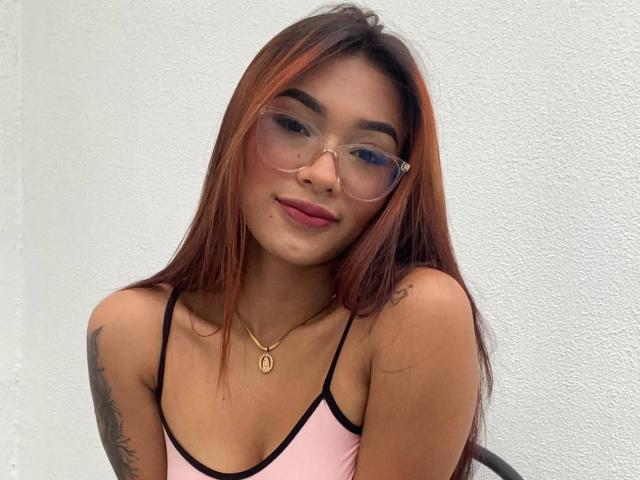SashaHans with long brown hair, glasses, pink top, tattoos, and a gold necklace, smiling slightly, showing her small tits and cute face.