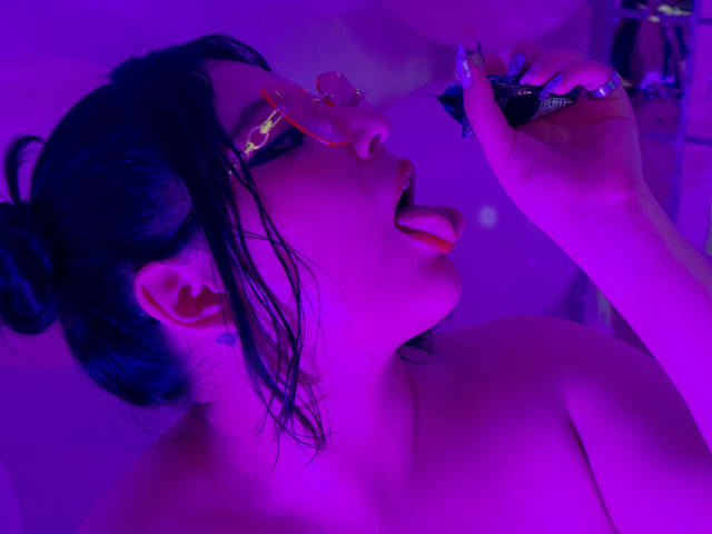 AnnaBeltrann, avec ses cheveux noirs attachés en chignon, éclairée en violet, la langue tirée, tenant un objet noir près de son nez, portant une bague jaune, nue, avec un tatouage visible sur le cou..
