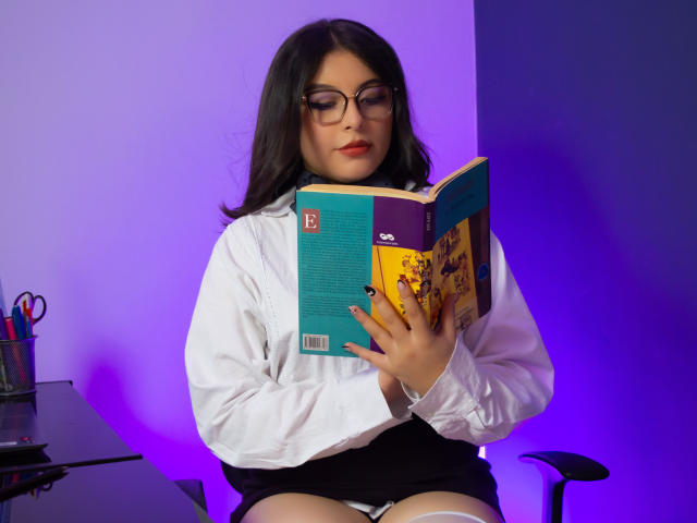 AnnaBeltrann, con gli occhiali e i capelli scuri, indossa una camicia bianca e una gonna nera, sta leggendo un libro con copertina viola e gialla, seduta su una sedia, con illuminazione viola..