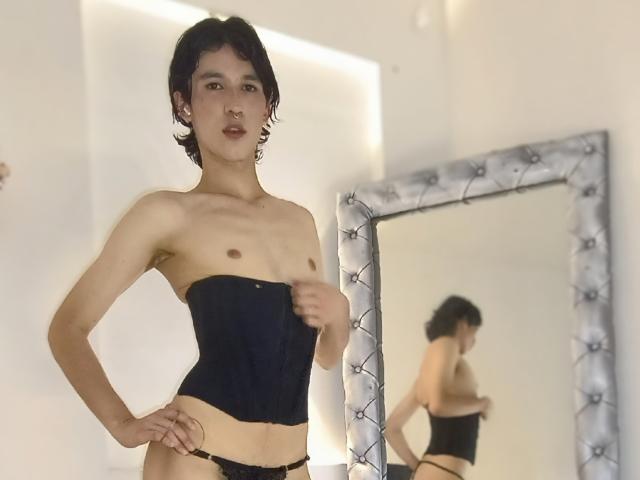 JullianXRicci with short black hair, wearing a black strapless corset and black panties, standing in front of a mirror showing their bare chest and reflection.