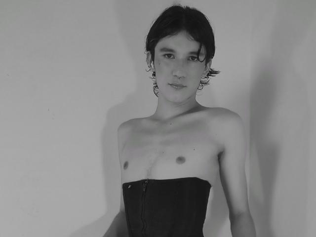 Black-and-white photo of JullianXRicci with wavy dark hair, wearing a strapless black corset, exposing small tits and a slim, flat-chested upper body. JullianXRicci has a neutral expression.