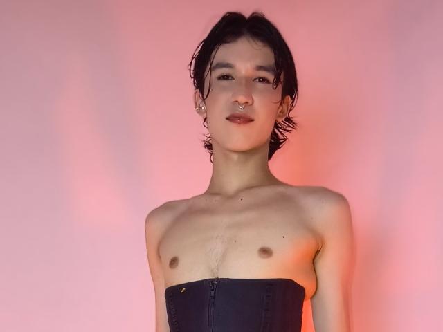 JullianXRicci, shirtless, with dark wavy hair, small nipples, and a black corset, standing against a pinkish background, looking slightly to the right with a subtle smile.