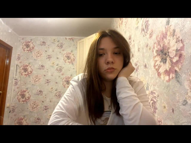AureliaEmswiler with brown hair, white shirt, and pensive expression, head resting on hand, in a floral-patterned room. Her tits are small, and she looks bored.
