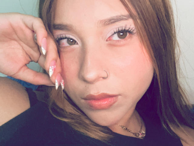HeraJuno has a cute face with light skin, brown eyes, and long brown hair. She's sporting a nose ring and has pink, white-manicured nails with a cute design.