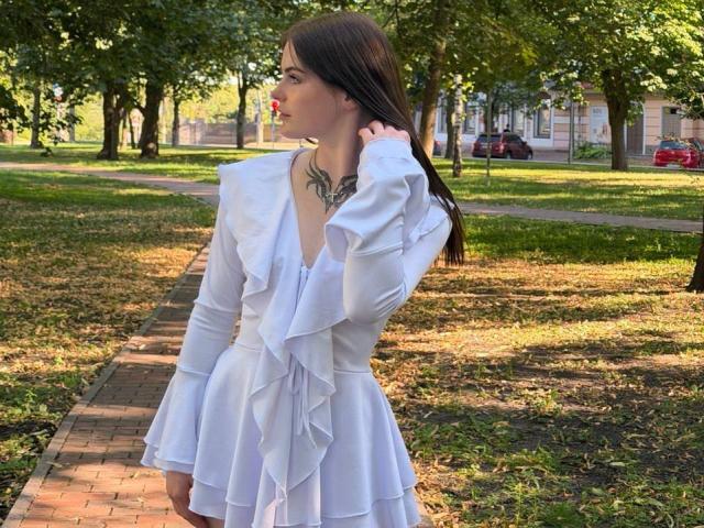 VirgieCrotts with long brown hair wearing a white ruffled dress, touching her hair, showing a tattoo on her neck, standing on a brick path in a sunny park.