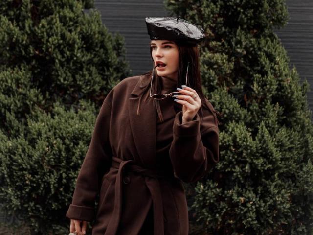 VirgieCrotts with brown coat, black hat, holding glasses, greenery in background, standing, looking to the side, slightly open mouth, dark green shrubs behind, hands up, relaxed pose.