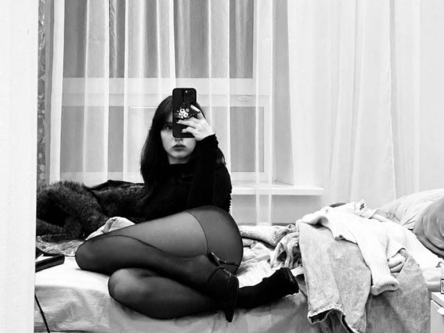 DanicaStuer with black hair, dark dress, and sheer black stockings, kneeling on a bed, taking a selfie with a phone, showing her ass and black heels.