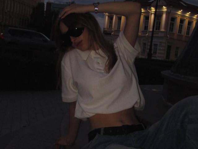 IsauraLatz with dark sunglasses, white shirt, and blue jeans, sitting with one arm raised, showing a flat stomach and black belt. Dimly lit, night setting.