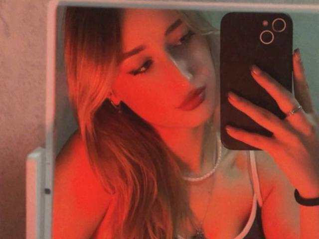 IsauraLatz with blonde hair, red-tinted face, wearing a white tank top and pearl necklace, taking a selfie with a black phone, showing her ring and bracelet.
