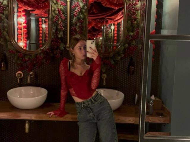 IsauraLatz with small tits, wearing a red lace top, denim pants, and blonde hair, taking a mirror selfie in a dark, ornate bathroom with red curtains.