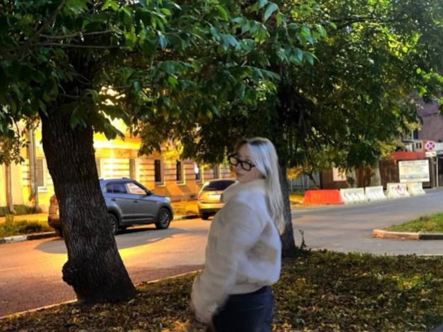 IsauraLatz stands in a park, facing sideways, wearing a fluffy white jacket, black glasses, and blue jeans, with long blonde hair, under a green-leaved tree.