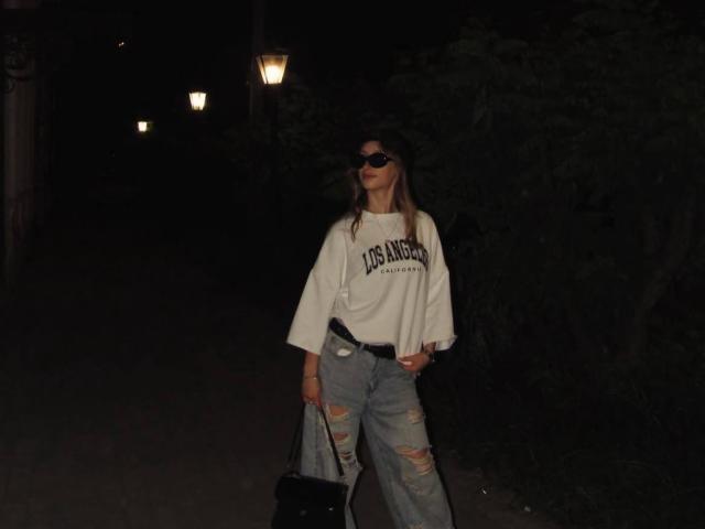 IsauraLatz stands in a dark night setting, wearing a white US Angels shirt, ripped blue jeans, black sunglasses, and holding a black bag, with street lamps in the background.