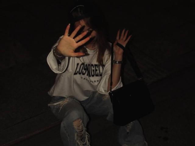 IsauraLatz with hands up, wearing a loose white "Los Angeles" shirt and ripped jeans, dark bag over shoulder, night, dim light, partially covering face.