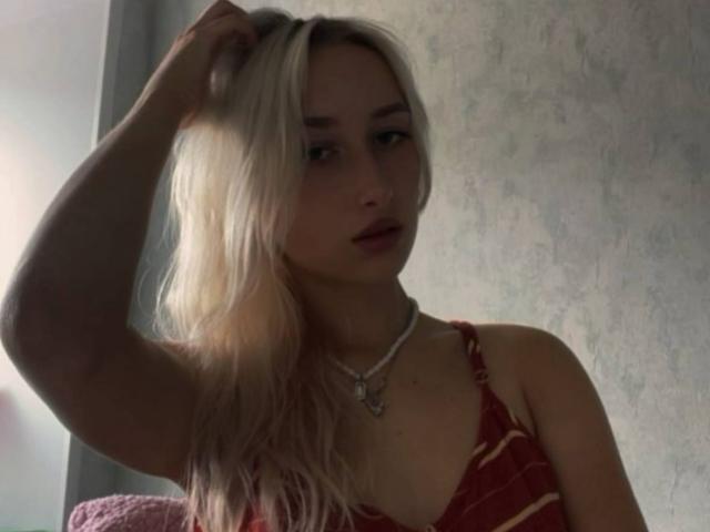 IsauraLatz with blonde hair, hand in hair, wearing a red striped top, necklace with heart pendant, soft light, slightly open mouth, looking down, subtle shadows on her face.