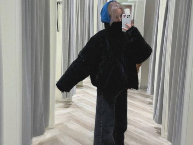 IsauraLatz with a blue cap, black fluffy coat, and dark jeans, taking a mirror selfie in a changing room, holding a phone with a pink case.