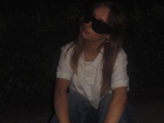 IsauraLatz with long brown hair, wearing a white shirt and blue jeans, sitting down, sporting big black sunglasses, looking to the side, in a dimly lit, dark background.