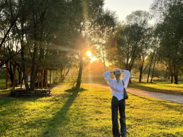 IsauraLatz stands in a grassy park with sun flares, hands behind head, wearing a white shirt, blue jeans, and a baseball cap, looking relaxed and confident.