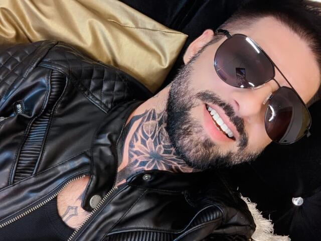 TommyDum with tattoos on his neck, black leather jacket, and sunglasses, smiling with a beard, chest tattoo visible, lying on a gold pillow, black background.