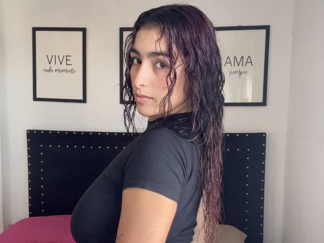 Rubyro with wet dark brown hair, black t-shirt, looking over shoulder, showing small nose boop, medium tits, in bedroom with black studded headboard, white wall.