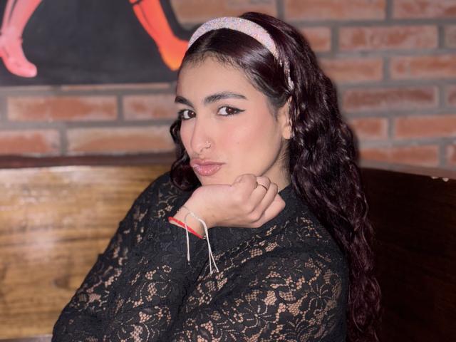 Rubyro with dark curly hair, wearing a lace black top and pink headband, pouting with nose ring, hand on chin, red bracelet, and white thread on wrist.