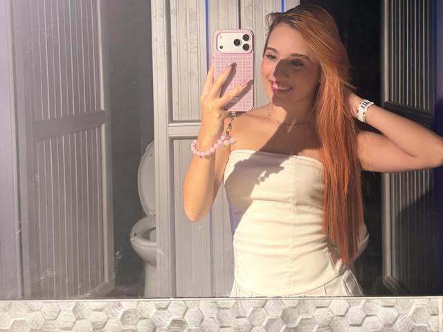 EmmaWat with long red hair, smiling, holding a pink phone, wearing a white strapless dress, showing her tits, in a mirror selfie, with a pink bracelet and watch.