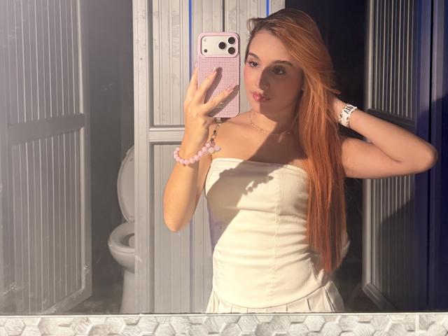 EmmaWat with long red hair, wearing a white strapless top, taking a mirror selfie, showing off her tits, pink phone case, pink bracelet, white watch, and pouty lips.