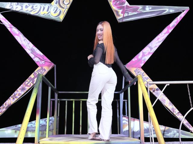 EmmaWat with long orange hair, black sheer top, white pants, standing on a colorful ride, looking back, smiling, showing her ass, black background, night, vibrant colors.