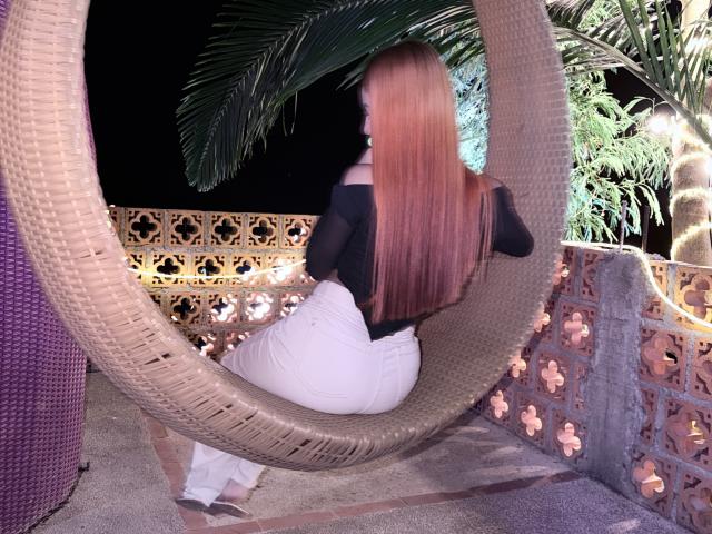 EmmaWat with long red hair, black off-shoulder top, and white pants, sitting in a woven hanging chair, showing her ass, in a night-time outdoor setting.