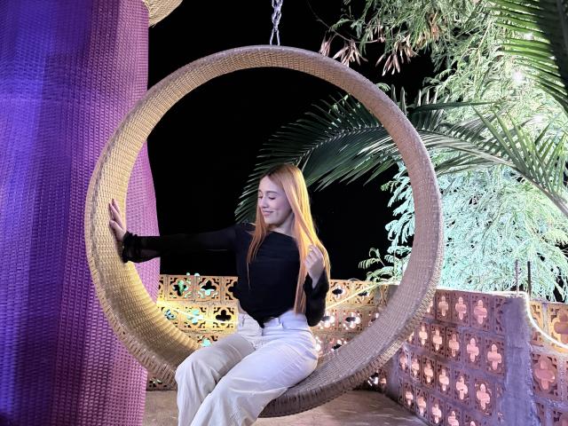 EmmaWat with long blonde hair, black top, white pants, sitting in a woven hanging circle chair, touching purple pillar, night time, palm leaves, calm expression, attractive, cute.