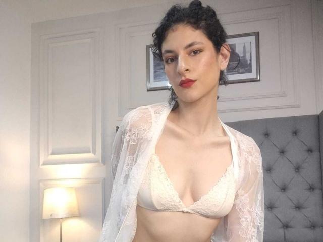 RoxyGraff with curly black hair, pale skin, and red lipstick, wearing a white lace bra and sheer robe, showing small tits and a flat ass, standing in a bedroom.
