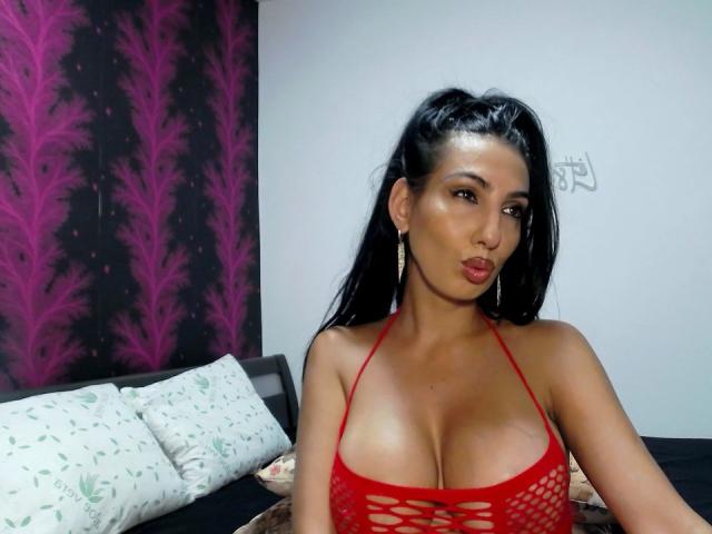 BrunetteBabe69 con grandi seni, indossa un top rosso con spalline e aperture, labbra carnose, lunghi capelli neri raccolti in una coda di cavallo e orecchini a cerchio dorati, seduta su un letto..