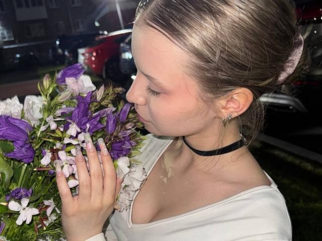 RaeleneRaya, avec ses seins légèrement visibles, portant un haut blanc et un collier noir, sentant des fleurs violettes et blanches, avec une petite pétale de fleur sur le nez..