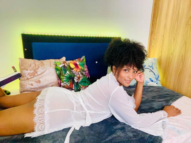 RoldanStefy, lying on a bed, wearing a white sheer lingerie dress, showing her curly afro, smiling at the camera, with her ass raised and her hand under her chin.