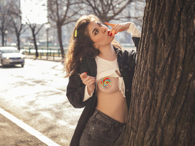 VanessaRaye steht neben einem Baum, hebt ihr Hemd, um eine farbenfrohe, wirbelnde Tätowierung auf ihrer Brust zu zeigen, lutscht an einem roten Lutscher und trägt einen schwarzen Mantel und Jeans..