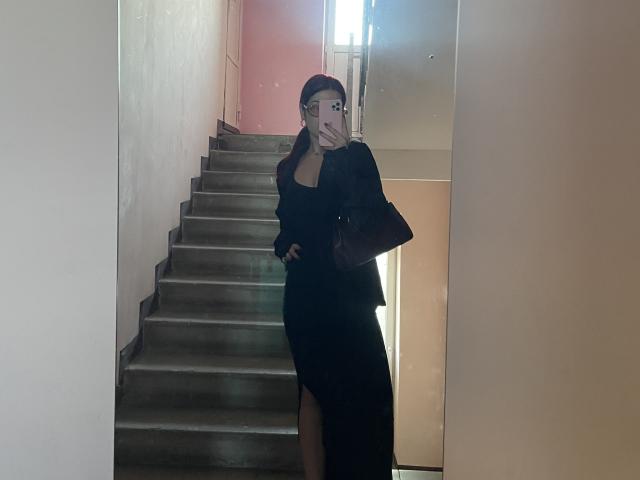 AlexXas in a black dress with a slit, holding a phone, standing on stairs, mirror selfie, hand on hip, brown bag, glasses, red hair, looking at phone.