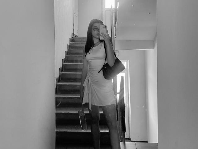 Black-and-white photo of AlexXas with long straight hair, glasses, holding phone to mouth, wearing dress, small ass, holding book, standing on stairs, bag on shoulder.