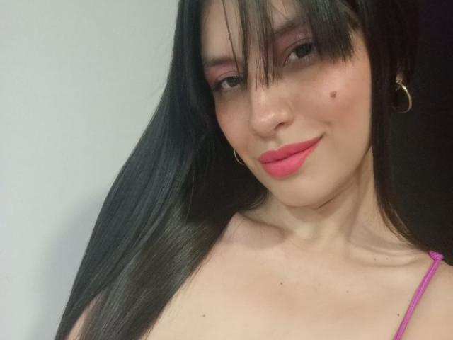 MadissonFontaine has long black hair, pink lipstick, and a small beauty mark on her right cheek. She's wearing a pink spaghetti strap top with a gold hoop earring.