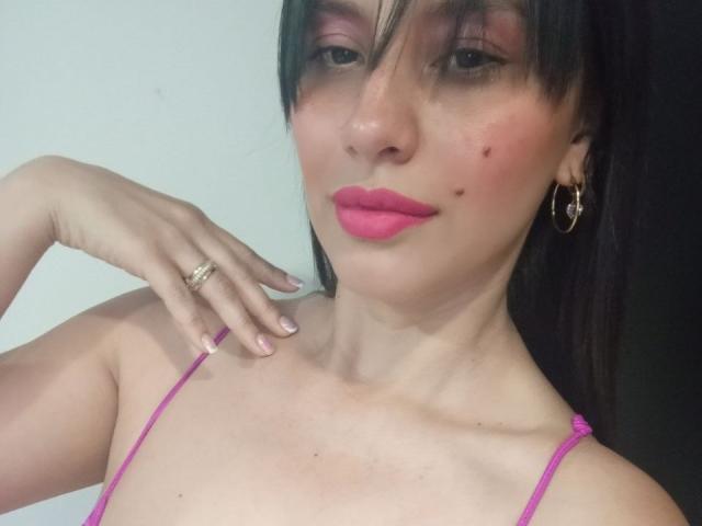 Madisson Fontaine, com batom rosa, cabelo preto, brincos de argola, um anel, alças finas rosa e algumas sardas no rosto, exibindo sua mão perto da clavícula..