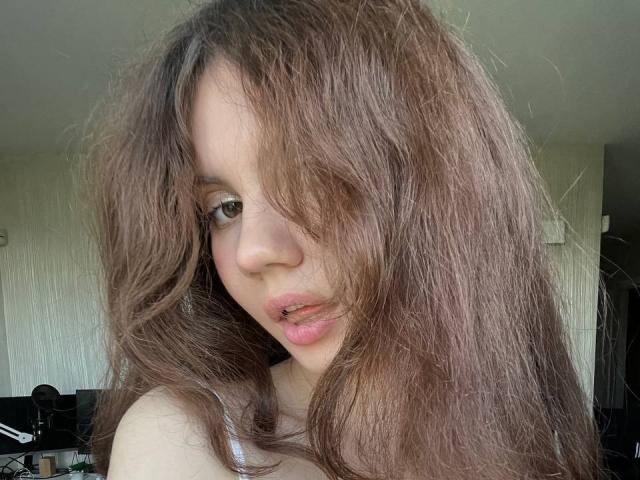 TanaFietek with long, messy brown hair covering one eye, slightly parted pink lips, and a subtle nose piercing, wearing a white top, looking at the camera.