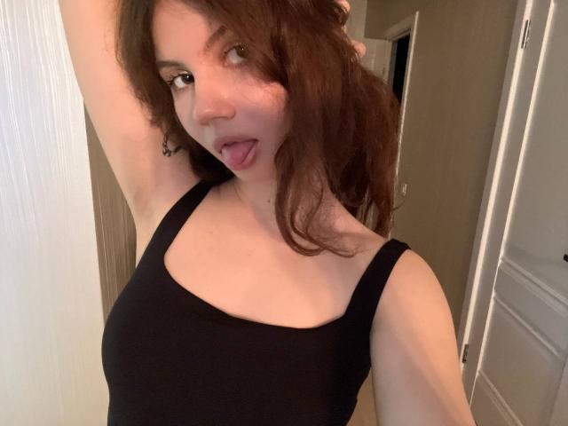 TanaFietek with wavy brown hair, black tank top, arm raised, tongue out, showing small tits, light skin, and a cute, playful expression in a selfie.