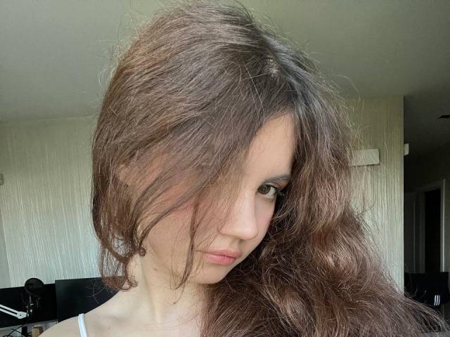 TanaFietek has messy brown hair covering one eye, pale skin, and a serious expression. She's wearing a white top, and her ass is not visible.
