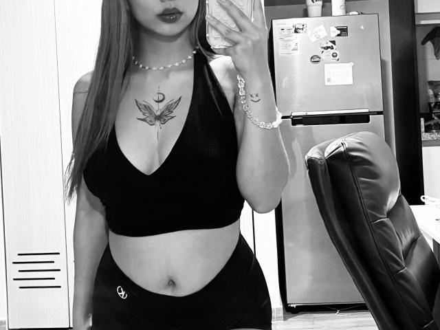 Samanta Carter hat langes Haar, ein schwarzes Crop-Top und schwarze Hosen, die ihr Schmetterlingstattoo auf der Brust zeigen. Sie trägt eine Perlenkette und ein Armband und hält ein Telefon..