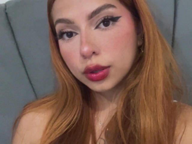 Samanta Carter hat roten Lippenstift, schwarzen Eyeliner im Winged-Stil und einen rosafarbenen Teint. Sie schaut in die Kamera mit voller Lippe, und ihr langes, rotes Haar fällt über ihre Schultern..