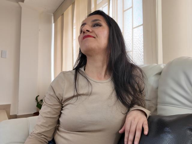 Cyntiia with long black hair, light skin, and red lipstick, wearing a beige long-sleeve shirt, sitting on a white leather couch, looking up with a slight smile.
