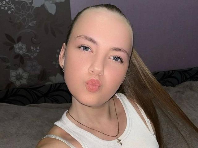 CherryMiss, de aspecto atractivo, con labios carnosos, ojos azules y cabello castaño largo, vestida con una camiseta blanca, lleva un collar y está en un selfie. Sus pechos son pequeños y está haciendo una mueca de beso..