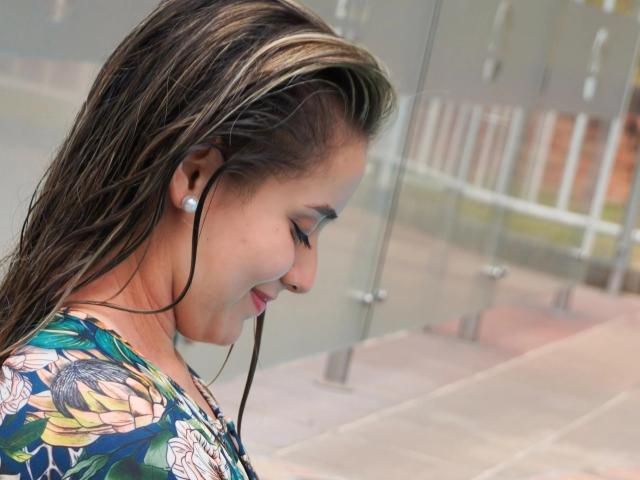 AngelaMonticellii with wet brown hair, wearing floral shirt, earbuds in, eyes closed, smiling, facing right, visible earrings, background blurred.